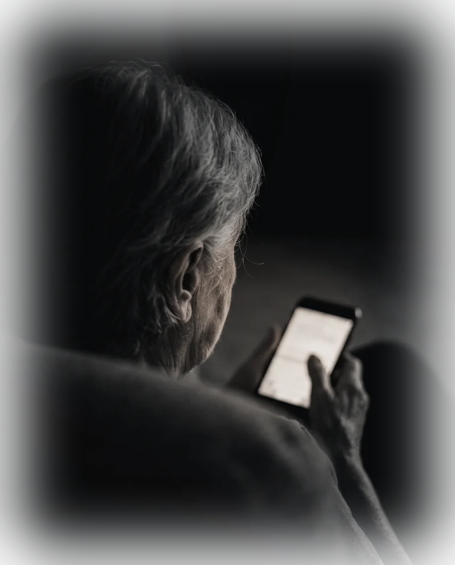 An older woman sitting alone in a dark room, reading a message on her phone. The screen is the only light.